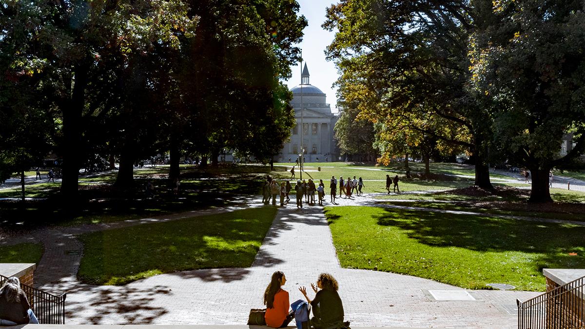 Main UNC quad during the day with students sitting on steps of South Building in foreground.